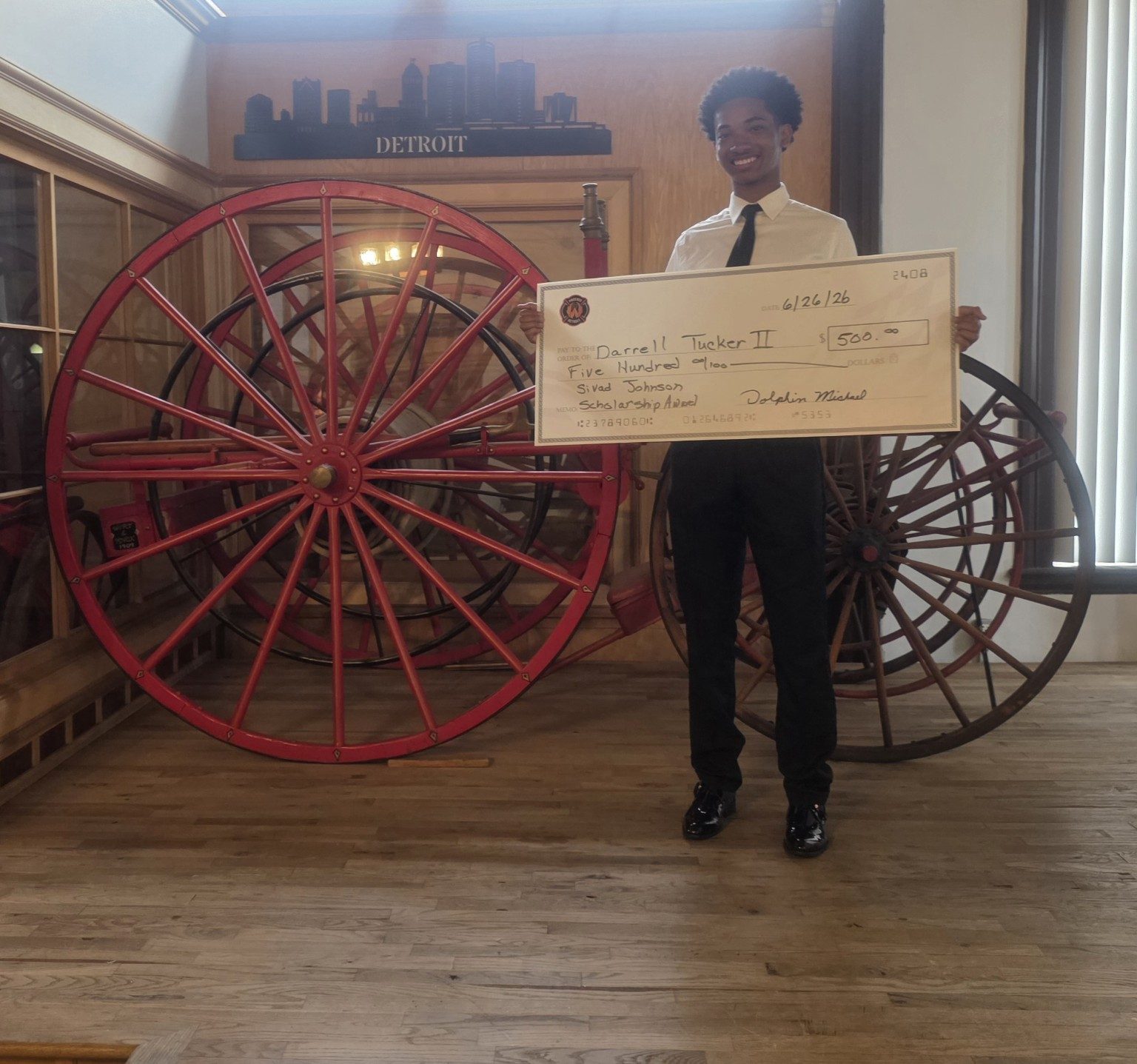 Darrell Tucker II, 2026 Sivad Johnson Scholarship recipient, receiving his scholarship check at Historic Engine 11 in front of the historic hose-reel carriage.